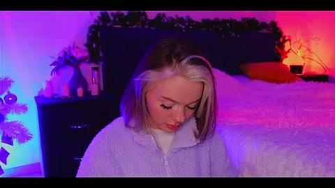 Media: Video of a young woman with blonde hair, wearing a light blue hoodie, sitting on a bed with white sheets. Background features a purple and orange lit room with holiday decorations.