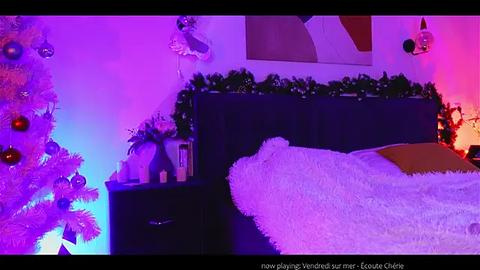 Media: Video of a cozy bedroom with a bed covered in a fluffy white blanket, purple and pink lighting, a decorated Christmas tree, and festive green garland on the wall.