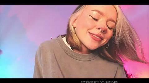 Media: Video of a smiling, fair-skinned, blonde-haired woman with long hair, wearing a grey sweater, against a vibrant purple and pink background. She has a nose ring and is looking down.