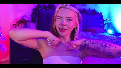 Media: A video of a smiling, tattooed, blonde woman with fair skin, wearing a strapless dress, lying on a bed under neon purple and blue lights, with a playful expression and one hand touching her chest.