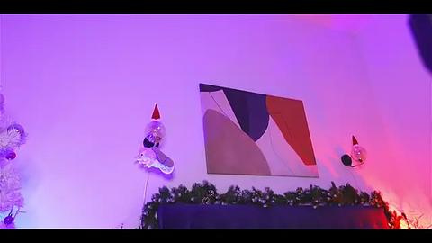 Media: Video of a festive indoor setting with a large abstract painting featuring geometric shapes and a garland of greenery. Two oversized, red and white elf figurines stand on either side, lit by purple and pink lights.