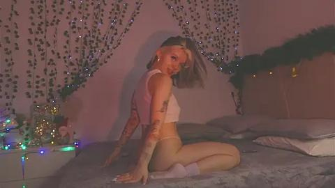 Media: Video of a blonde, tattooed woman in white lingerie, kneeling on a bed with gray sheets, surrounded by fairy lights and green vines.