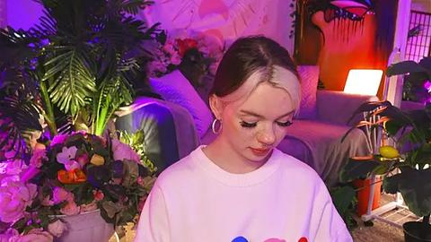 Media: Video of a young woman with light skin, dark brown hair, and hoop earrings, wearing a white T-shirt, standing among a lush indoor garden with purple lighting, flowers, and plants, creating a whimsical, serene atmosphere.