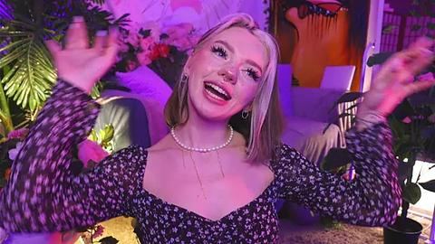 Media: Video of a smiling, fair-skinned blonde woman in a black floral dress, wearing a pearl necklace, with arms raised in excitement. Background features vibrant potted plants, hanging decor, and soft lighting.