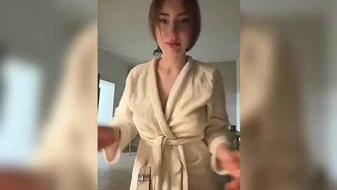 Media: Video of a young Caucasian woman with short brown hair, wearing a beige bathrobe, holding a phone in a dimly lit, neutral-toned room.