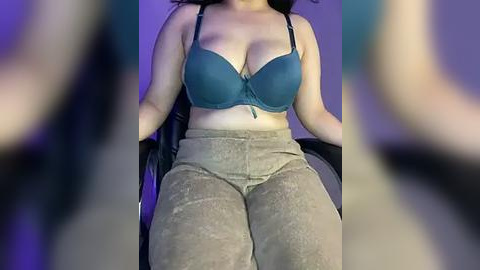 Media: A video of a woman with medium skin tone and long dark hair, wearing a teal bra and loose beige pants, sitting in a black chair. The background is blurred with purple lighting.