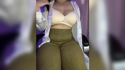 Media: A video of a light-skinned woman with a curvy figure, wearing a white blouse over a beige bra and high-waisted olive-green pants, seated in a dimly lit room.