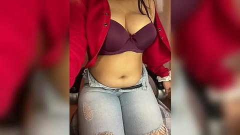 Media: Video of a woman with medium skin tone, wearing a purple bra, light blue distressed jeans, and a red cardigan, sitting on a chair.