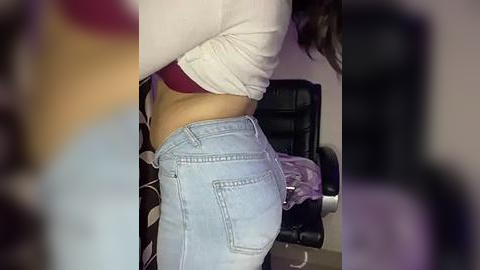 Media: Video of a woman in a white long-sleeve top and light blue jeans, showing her toned midriff and buttocks. Background includes a black leather chair and purple fabric.