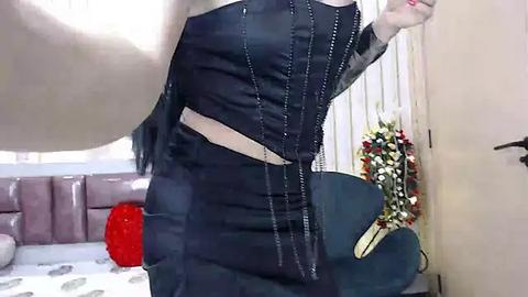 Media: Video of a woman with pale skin, wearing a tight, black, corset-style top and matching skirt, standing in a room with beige walls, a tufted couch, and a red balloon.