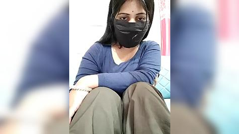 Media: Video of a woman with long black hair and a black face mask, wearing a blue long-sleeve top and gray pants, sitting indoors with a blurred background.