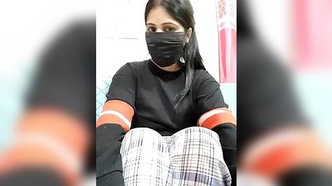 Media: A video of a woman with long black hair, wearing a black mask, black shirt, and plaid pants, sitting in a room with a blurred background.