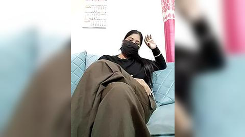 Media: Video of an Asian woman with long black hair, wearing a black mask, black top, and brown blanket, sitting on a light blue couch, with blurred figures in the foreground.