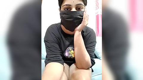 Media: Video of a South Asian woman with short hair, wearing a black mask, black t-shirt, and grey shorts, sitting on a light blue couch. The background features a white wall with pink and red patterns.