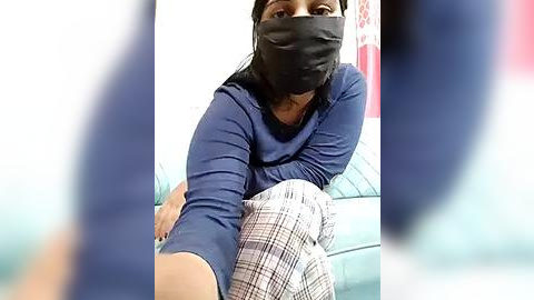 Media: Video of a woman with dark skin, wearing a black mask, blue shirt, and plaid pants, sitting on a teal sofa in a room with white walls and red decor.