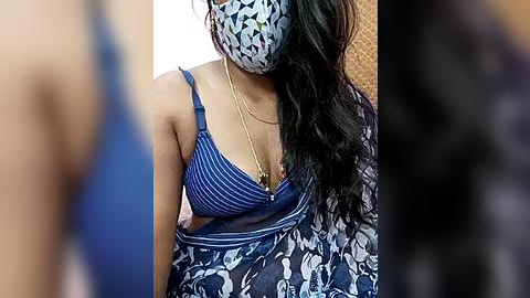 Media: Video of a woman with medium brown skin wearing a blue and white floral-patterned dress, a blue bra, and a blue and white patterned face mask, sitting indoors with a blurred background.