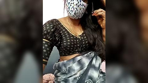 Media: Video of a woman in a black and white saree with a floral pattern, wearing a white mask with black polka dots, adjusting her hair with a hand, indoors.