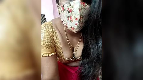 Media: Video of a woman with a floral mask, wearing a yellow textured top and red sari, her long black hair partially visible, sitting indoors.