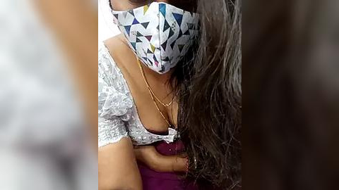 Media: Video of a woman with medium brown skin, wearing a white mask with geometric patterns, a lacy white blouse, and a purple skirt. Her long, wavy black hair partially obscures her face.