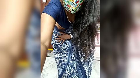 Media: A video shows a woman in a blue and white sari bending over, wearing a colorful mask, in a blurred, possibly urban setting.