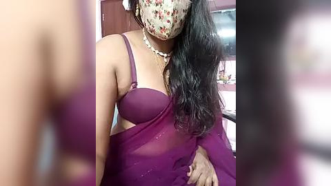 Media: A video of a South Asian woman wearing a purple sari, floral mask, and pearl necklace, with blurred background.