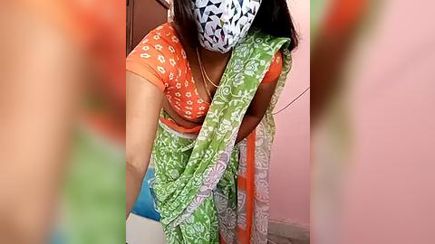 Media: Video of a woman in a green sari with white floral patterns, wearing an orange top with white floral patterns and a blue face mask, leaning forward in a pink room.