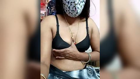 Media: A video of a woman wearing a black bra and a face mask, covering her mouth, with a tie-dye skirt, sitting indoors, blurring the background.