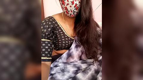 Media: Video of a woman wearing a black saree with white floral patterns, a red face mask, and a gold necklace. Background is blurred, with a pink wall visible.