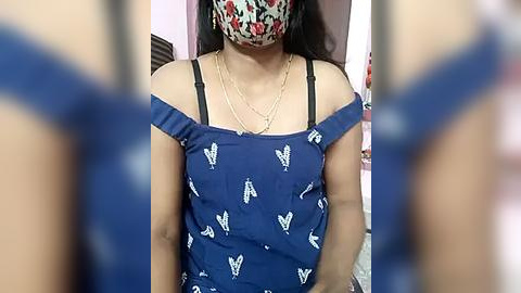 Media: Video of a woman wearing a floral-patterned mask, navy blue off-shoulder dress with white butterfly prints, and a gold necklace.