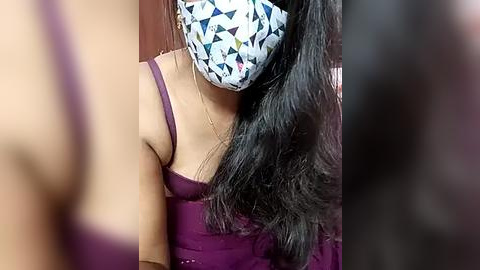 Media: Video of a woman with long black hair, wearing a purple dress, a face mask with geometric patterns, and a gold necklace.