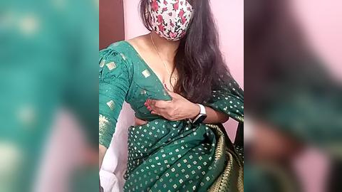 Media: Video of a woman in a green sari with white polka dots, wearing a floral face mask, holding her chest, with a blurred background.