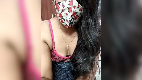 Media: Video of a South Asian woman with long, wavy black hair, wearing a pink lace bra, floral face mask, and blue-patterned dress. Blurry background suggests indoor setting.