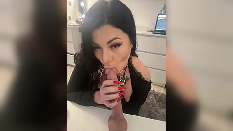 Media: A video of a woman with long black hair, fair skin, and full lips, wearing a black off-shoulder top, performing oral sex on a large, erect penis in a modern kitchen with white cabinets and a computer monitor in the background.