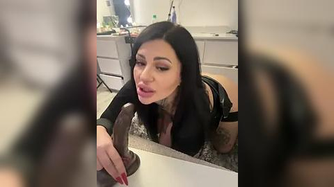 Media: Video of a tattooed woman with long black hair, wearing a black outfit, licking a dildo on a kitchen counter in a modern kitchen with white cabinets and a grey backsplash.