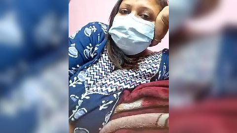 Media: A video of a South Asian woman with medium brown skin, wearing a light blue face mask, blue patterned top, and red blanket, resting her head on her hand, against a pink background.