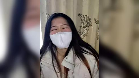 Media: A video of an Asian woman with long black hair, wearing a white mask, closed eyes, and a light beige jacket, standing against a beige curtain with floral patterns.