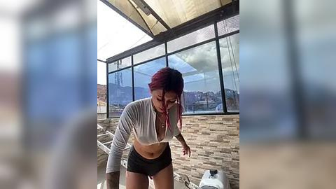 Media: Video of a young woman with red hair, wearing a grey crop top and black shorts, bending over on a sunlit balcony with a view of mountains.