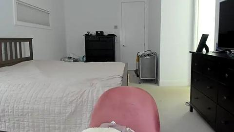 Media: Video of a minimalist, modern bedroom with a large, neatly made bed in the center, a black dresser on the right, and a pink chair in the foreground.