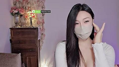 Media: Video of an East Asian woman with long black hair, wearing a white mask, white cardigan, and a pink top, in a room with floral wallpaper, a wooden dresser, and a glowing lamp.