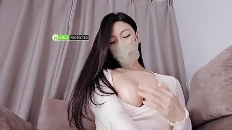 Media: Video of an East Asian woman with long black hair, wearing a beige face mask, a white top, and revealing her large breasts. She's seated on a beige couch, with light grey curtains in the background.