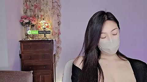 Media: Video of a woman with long black hair, wearing a face mask, a black dress, and a white top, standing in a room with a wooden dresser, a lamp, and a floral backdrop.