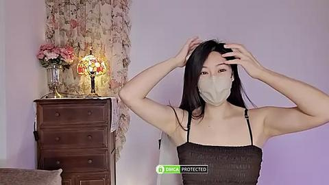 Media: Video of an East Asian woman with long black hair, wearing a face mask, adjusting her hair in a bedroom with a floral-patterned wall, wooden dresser, and a glowing lamp.