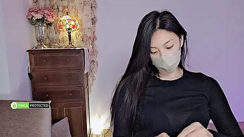 Media: Video of an Asian woman with long black hair and a surgical mask, wearing a black long-sleeved top, sitting in a room with a wooden dresser, a floral-patterned wall, and a lamp.