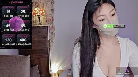 Media: A video of a young Asian woman with long black hair, wearing a white top and mask, sitting indoors. Background includes a vintage lamp, wooden dresser, and digital weather display.