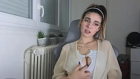 Media: Video of a young woman with fair skin, glasses, and a high ponytail, wearing a sheer top and beige cardigan, sitting in a grey office chair. Background shows a white radiator, potted plant, and a black helmet.