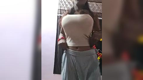 Media: Video of a woman with medium brown skin, wearing a beige tube top and gray skirt, standing indoors, with a lace curtain background.