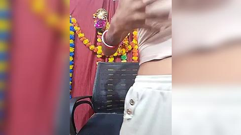 Media: A video of a person with fair skin, wearing a pink shirt, white shorts, and a red sari adorned with a colorful garland, standing in front of a blackboard with a list written in white.