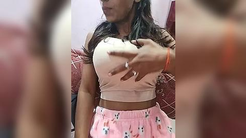 Media: Video of a South Asian woman with medium skin tone, long dark hair, wearing a tight, light pink crop top and pink pajama pants with bunny prints. She touches her chest. Background includes a red sofa with a patterned cushion.