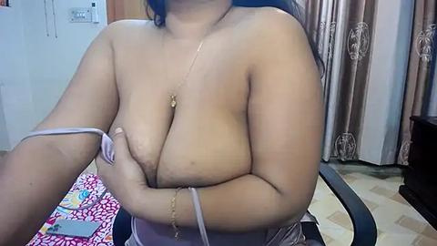 Media: A video of a topless Asian woman with medium-sized breasts, wearing a light pink dress and a gold necklace, sitting in a room with beige walls, curtains, and a colorful patterned blanket.