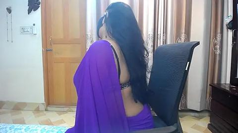 Media: Video of a woman with long black hair, wearing a purple saree and black bra, sitting on a black office chair in a modern, tiled bedroom with wooden doors and patterned curtains.